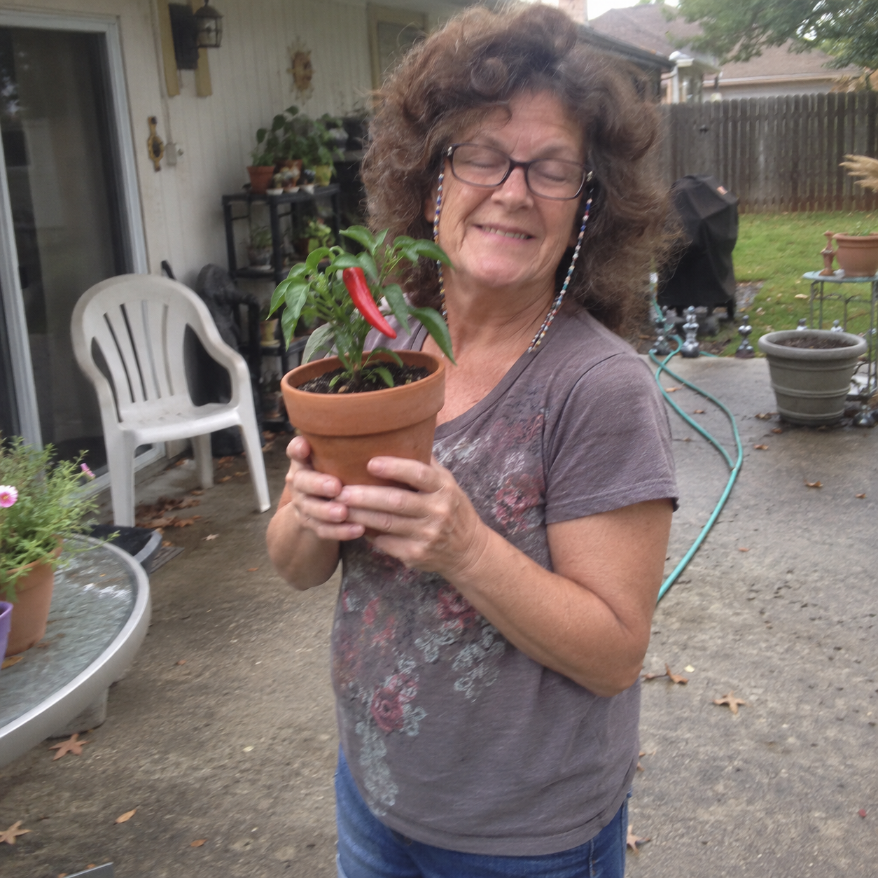 Brenda, smiling with her eyes closed, holds a terracotta pot with a small pepper plant bearing one red chili pepper.